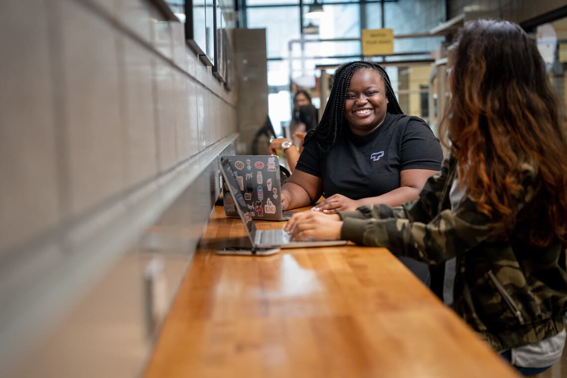Two students sit at a counter height wooden table, each with a laptop. They are smiling at each other int he UTSC Library.