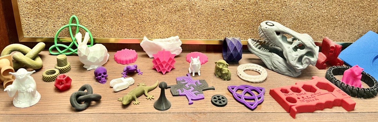 A display of colourful projects created on a 3D Printer is spread across a table, including a Yoda, snakes, lizards, a dinosaur skull and a bunny rabbit.