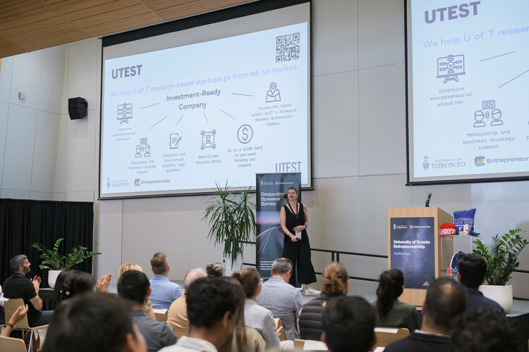 A white woman in a black dress addresses a crowd on a stage at U of T's Accelerator Fest. A slide show projected behind her shares information about a new web app.