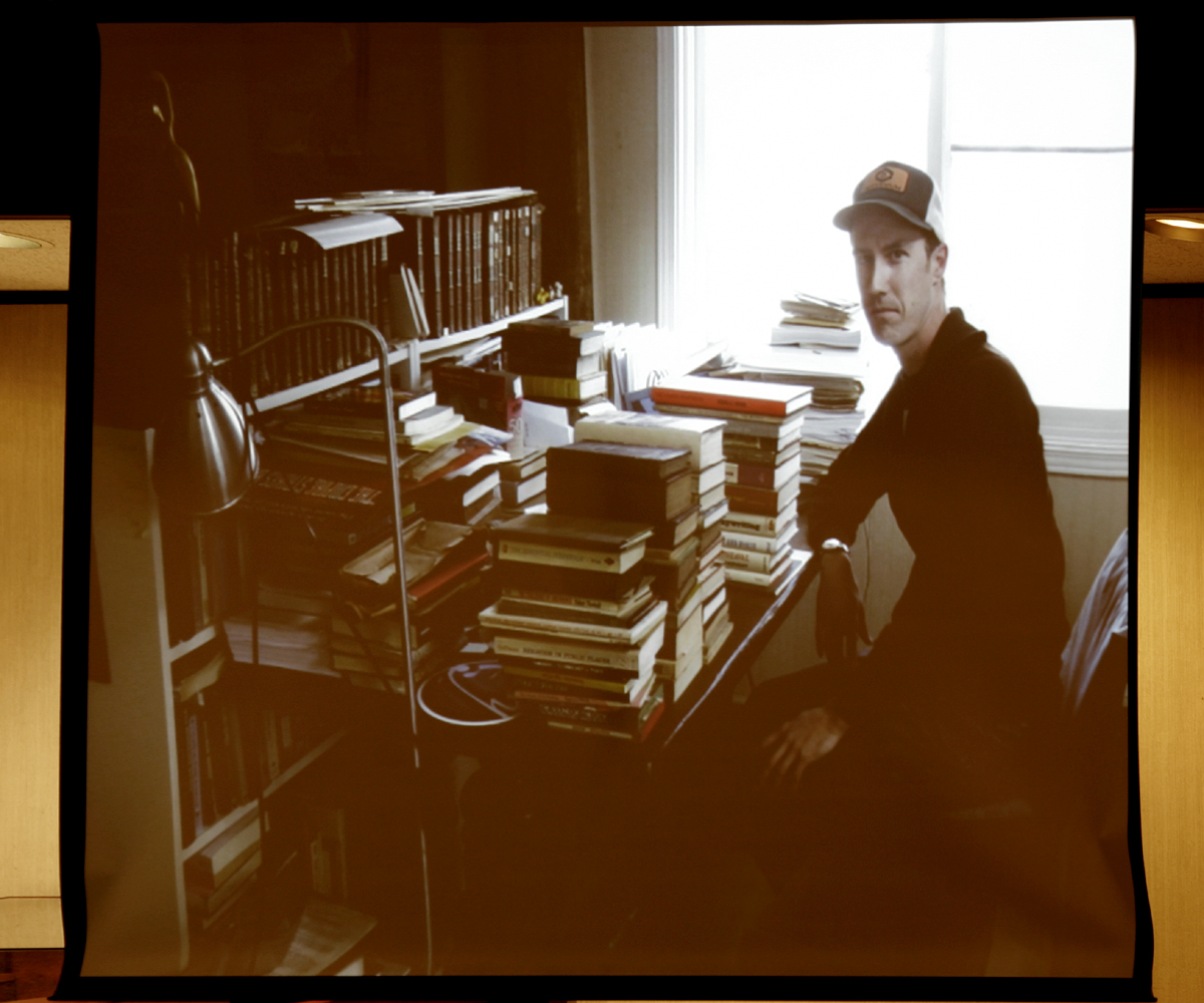 A white man in his 30s wearing a baseball cap sits at a desk in front of a stack of books.