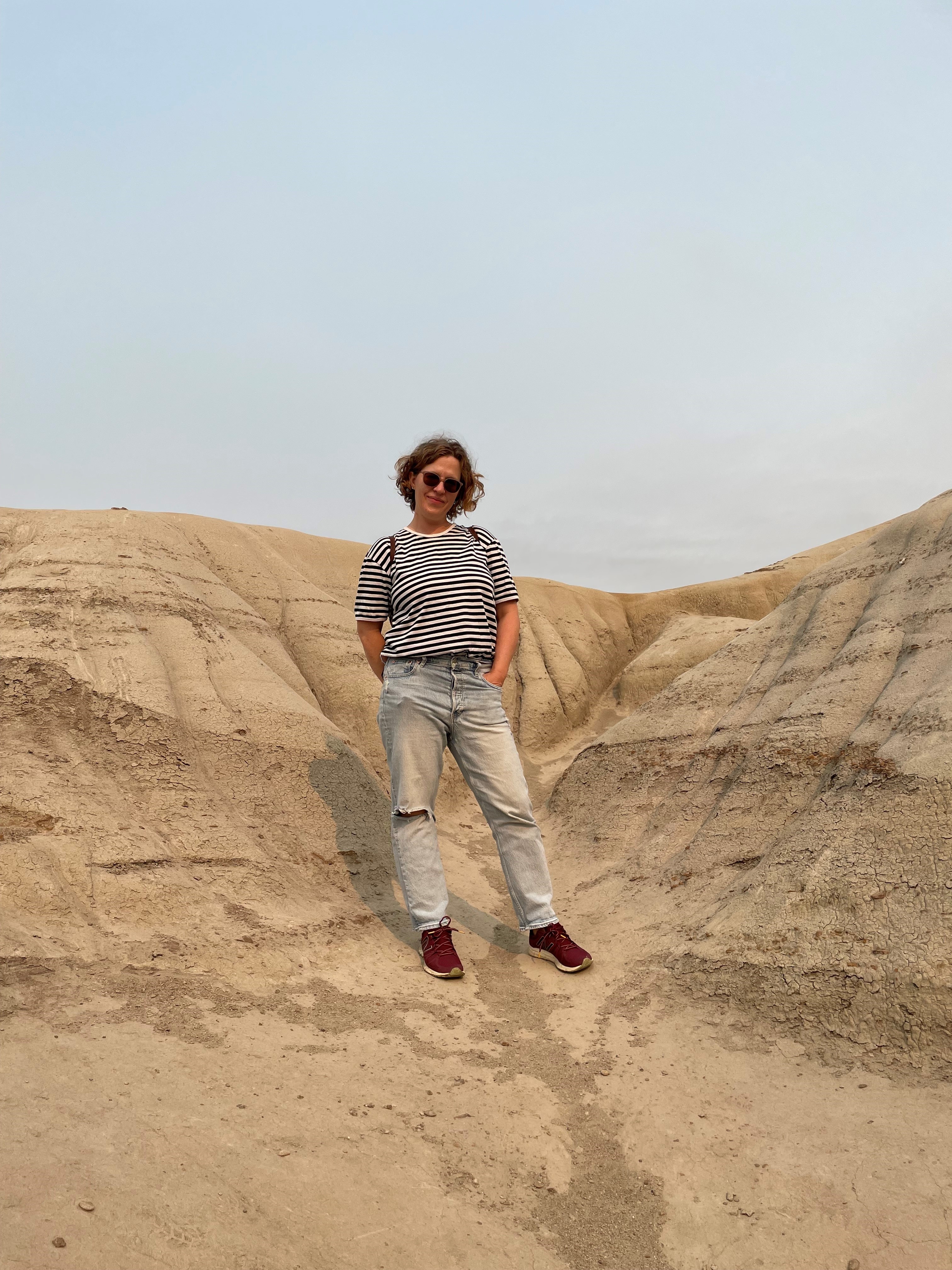 A white woman with short dark hair, a striped shirt and blue jeans stands on a mountain in Drumheller, Alberta.