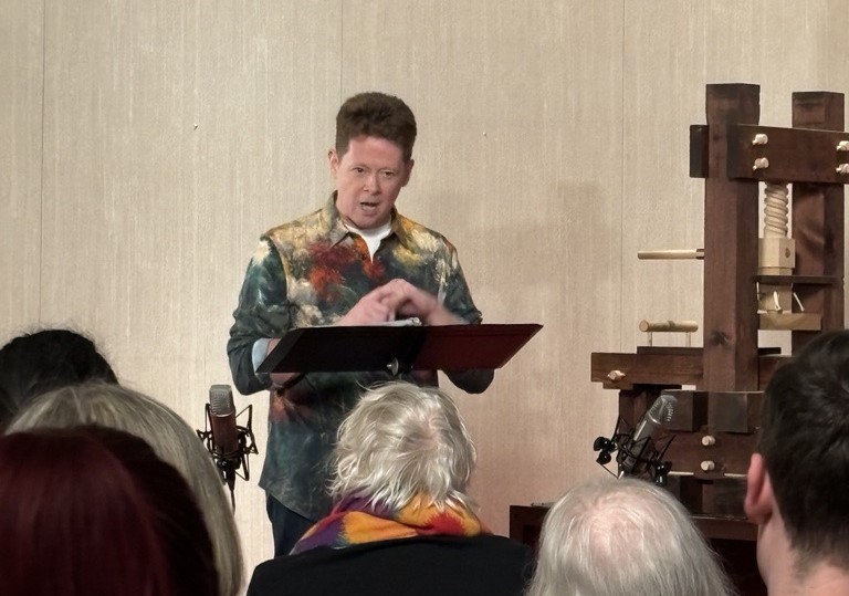 A white man in a patterned shirt and red hair standing at a podium and reading aloud from a copy of James Joyce's Finnegan's Wake. An expression of intensity is on his face and his hands are clasped in front of him.