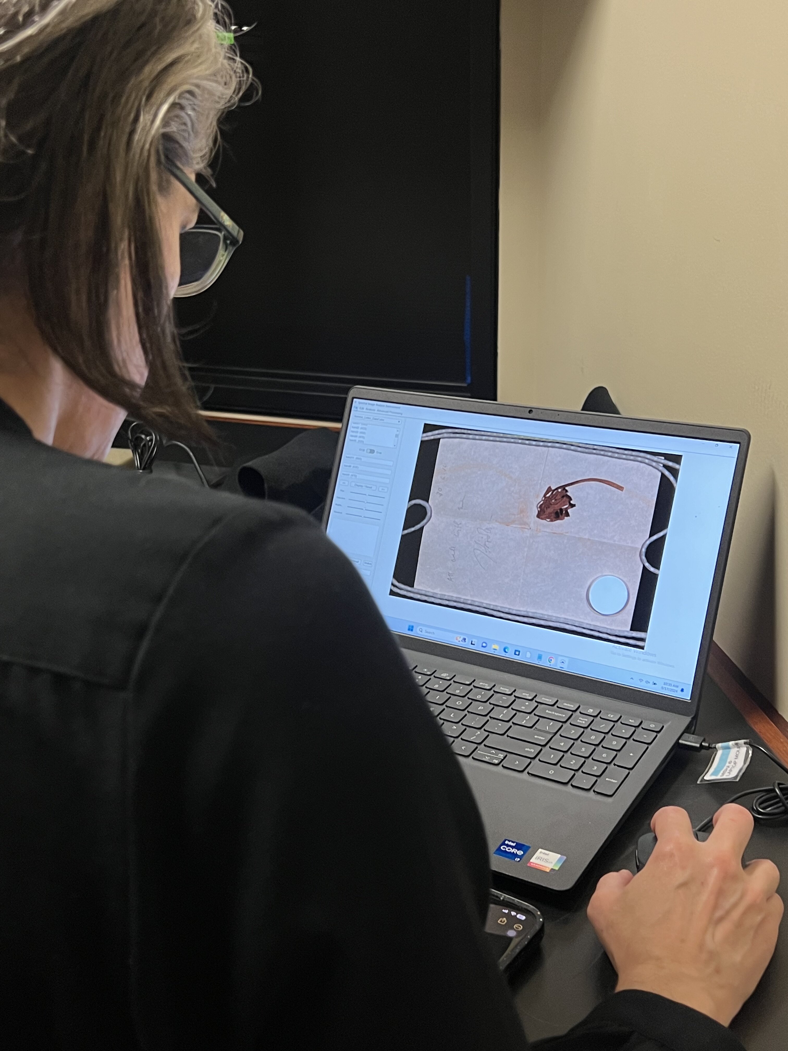 A woman in a black blouse scans a flower and letter on a multi spectral imaging system and analyzes results on the computer.