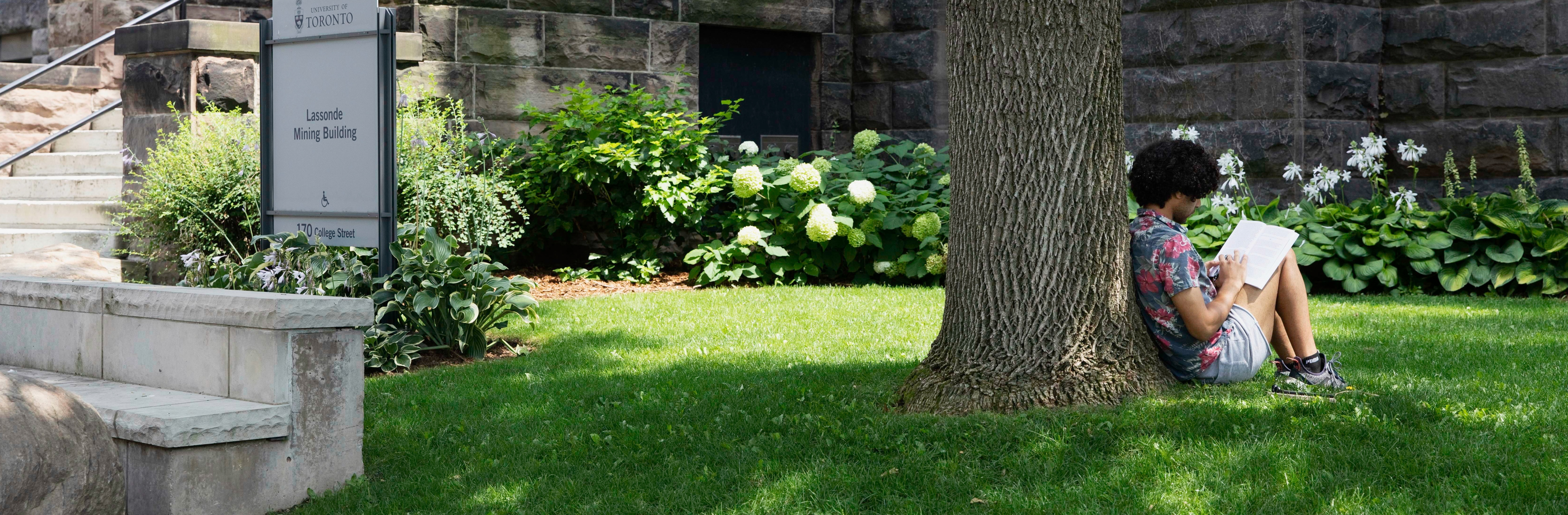 person sits reading against a tree on a field outside of a building on a sunny day