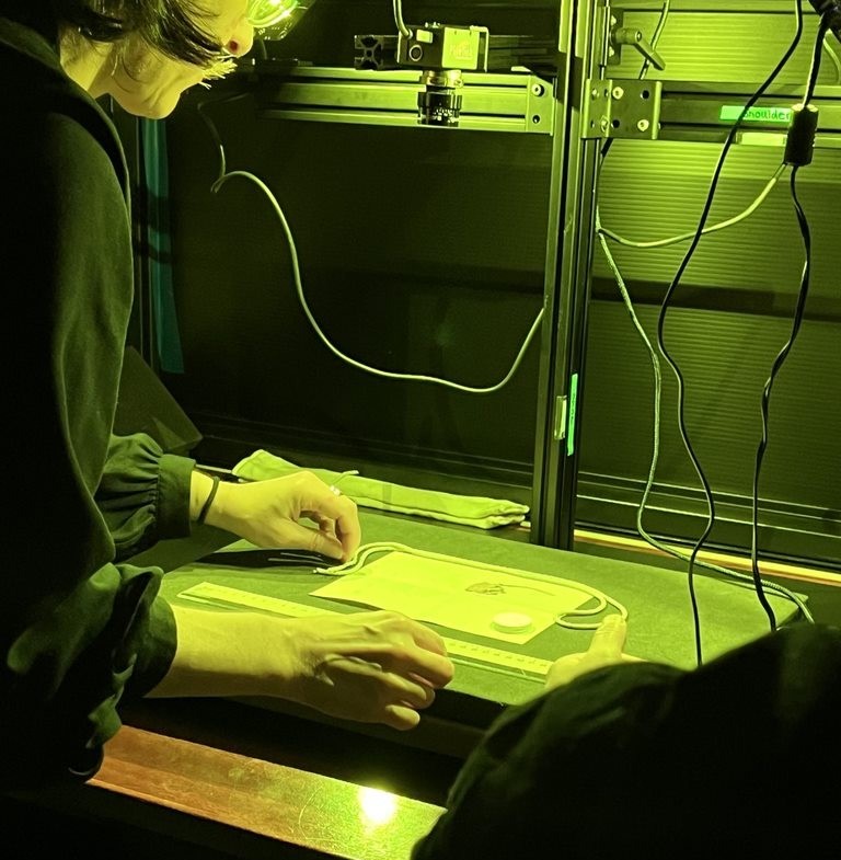 A white woman in a black blouse places a letter and dried flower under a scanning device to try and identify what type of flower it is. The room is lit in green to assist with the scanning.