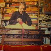 A Tibetan man with grey hair opens a traditional Tibetan text wrapped in beige cloth with red tie. A stack of colourful texts is piled behind him.