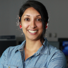 Women with brown skin complexion, dark hair up with a big smile wearing red earrings and blue jean shirt