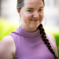 a white woman with brown hair in a braid and large thin hoop earrings, wearing a lavender top