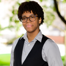 A photo of a person wearing glasses and a vest from the chest up. Background is blurred.
