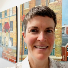 A person with short brown hair, pinkish skin, and earrings, standing in front of two paintings.