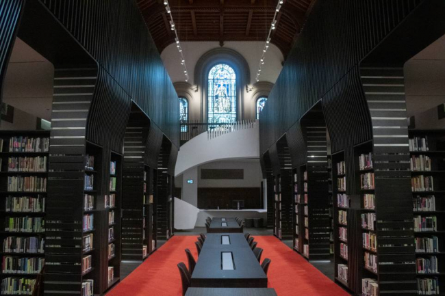 Photo of the University College library featuring spiral staircase, stained glass window, bookstacks, and large tba